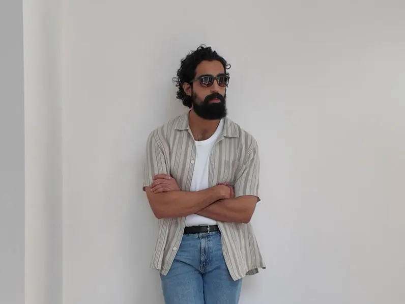 Tom Cheema wearing a striped shirt over a vest with blue jeans, a belt and sunglasses