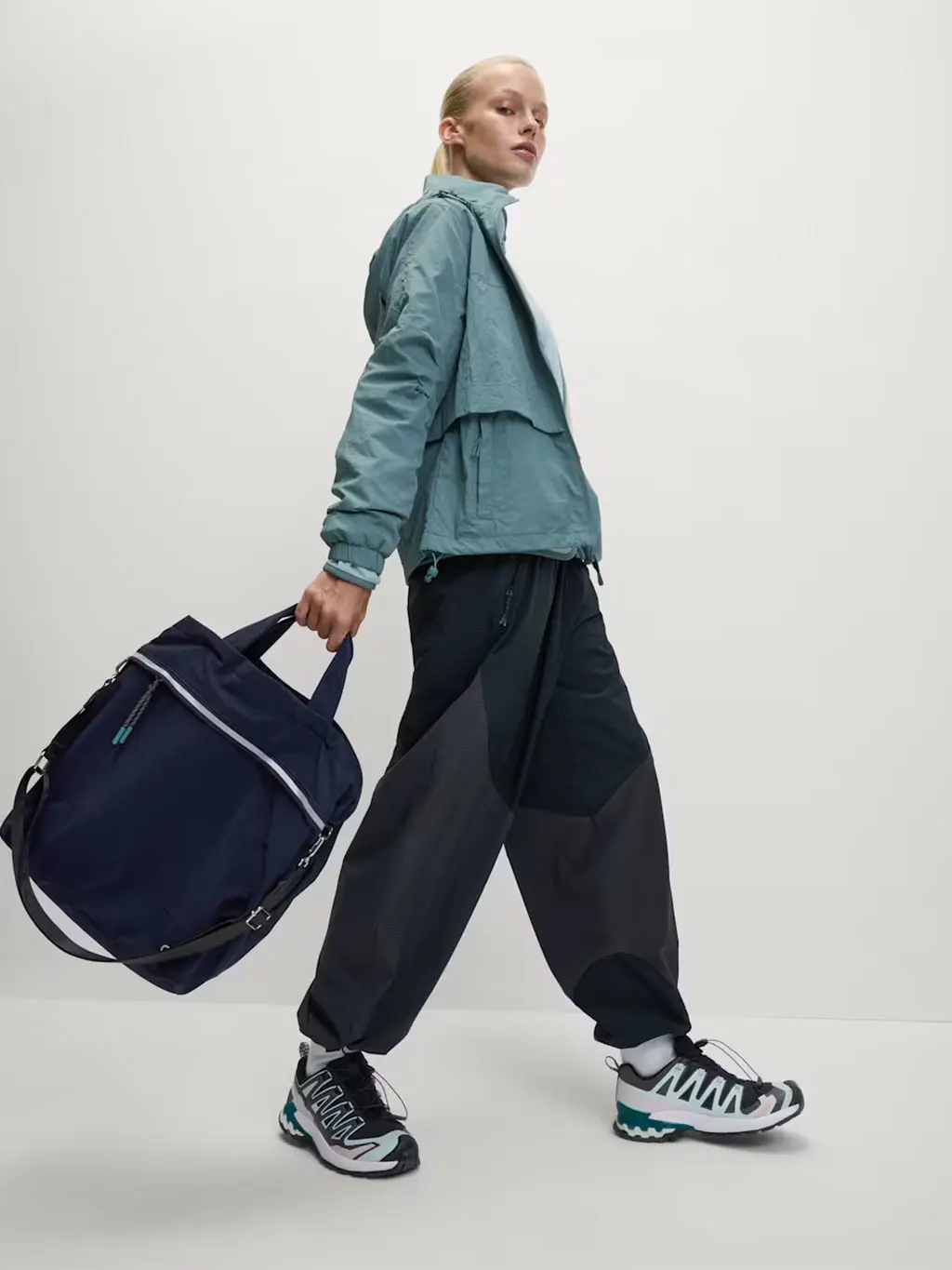 Woman wearing a blue waterproof jacket and trousers with trainers and a navy tote bag.