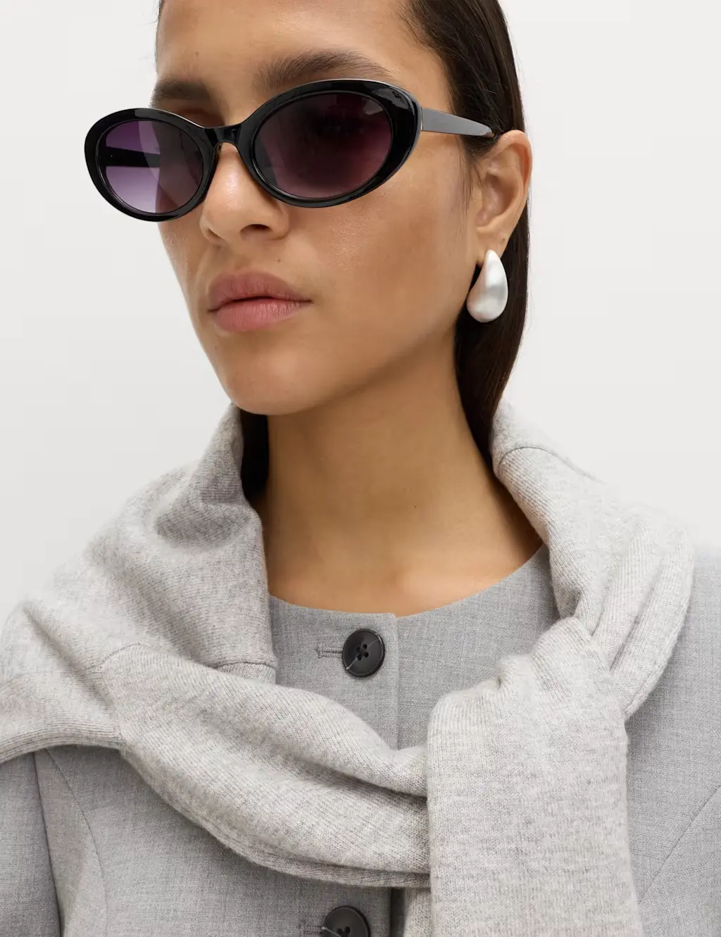 A woman wears a pair of black sunglasses, a grey knotted sweater and a collarless blazer