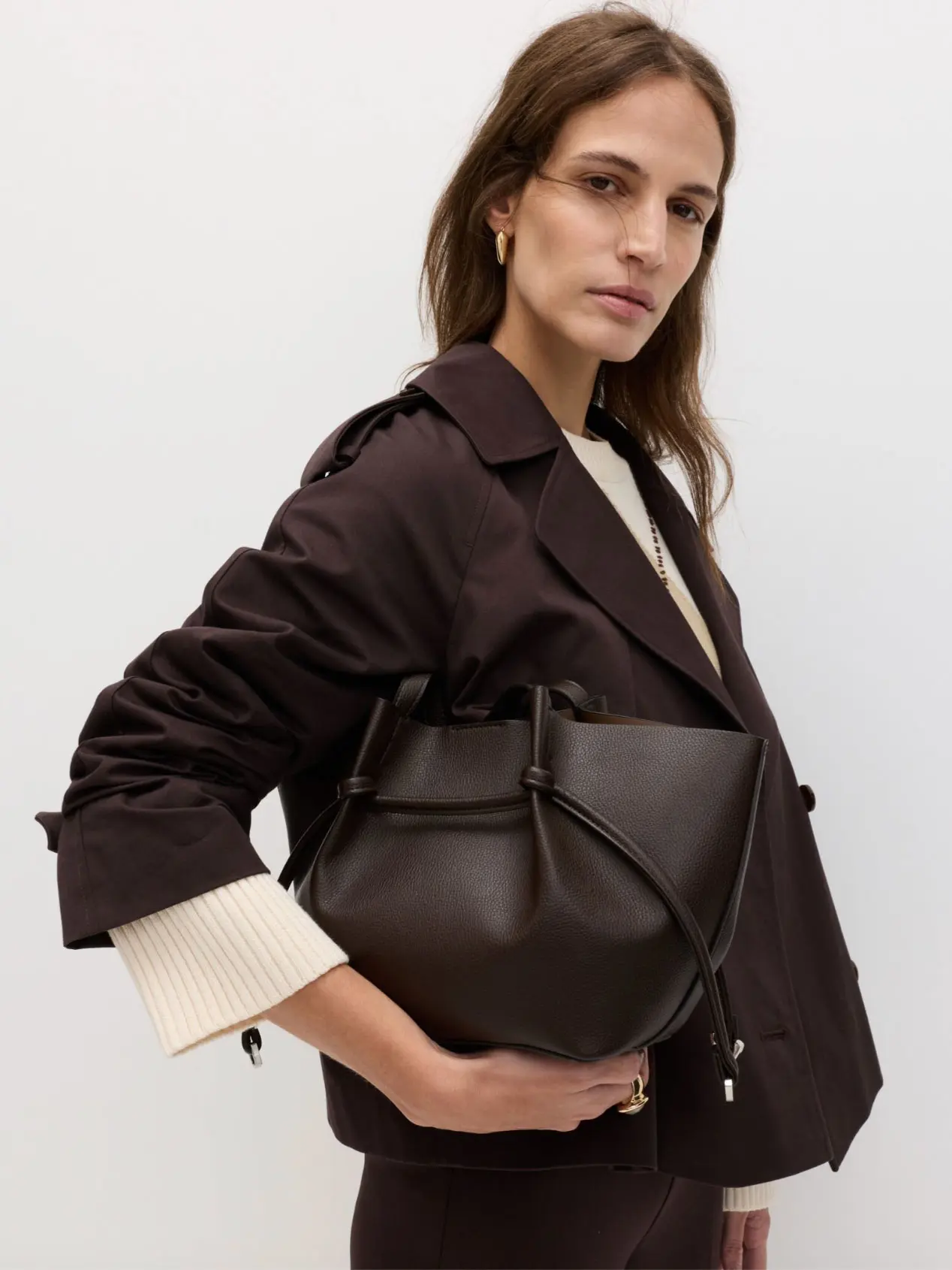 Woman wearing brown cropped trench coat over neutral jumper, holding brown leather bag
