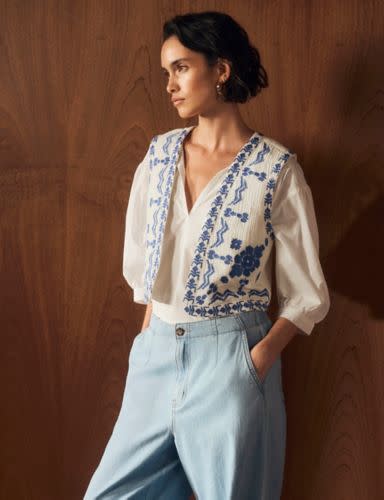 Woman wearing blue and white embroidered short gilet with blue trousers. Shop Per Una