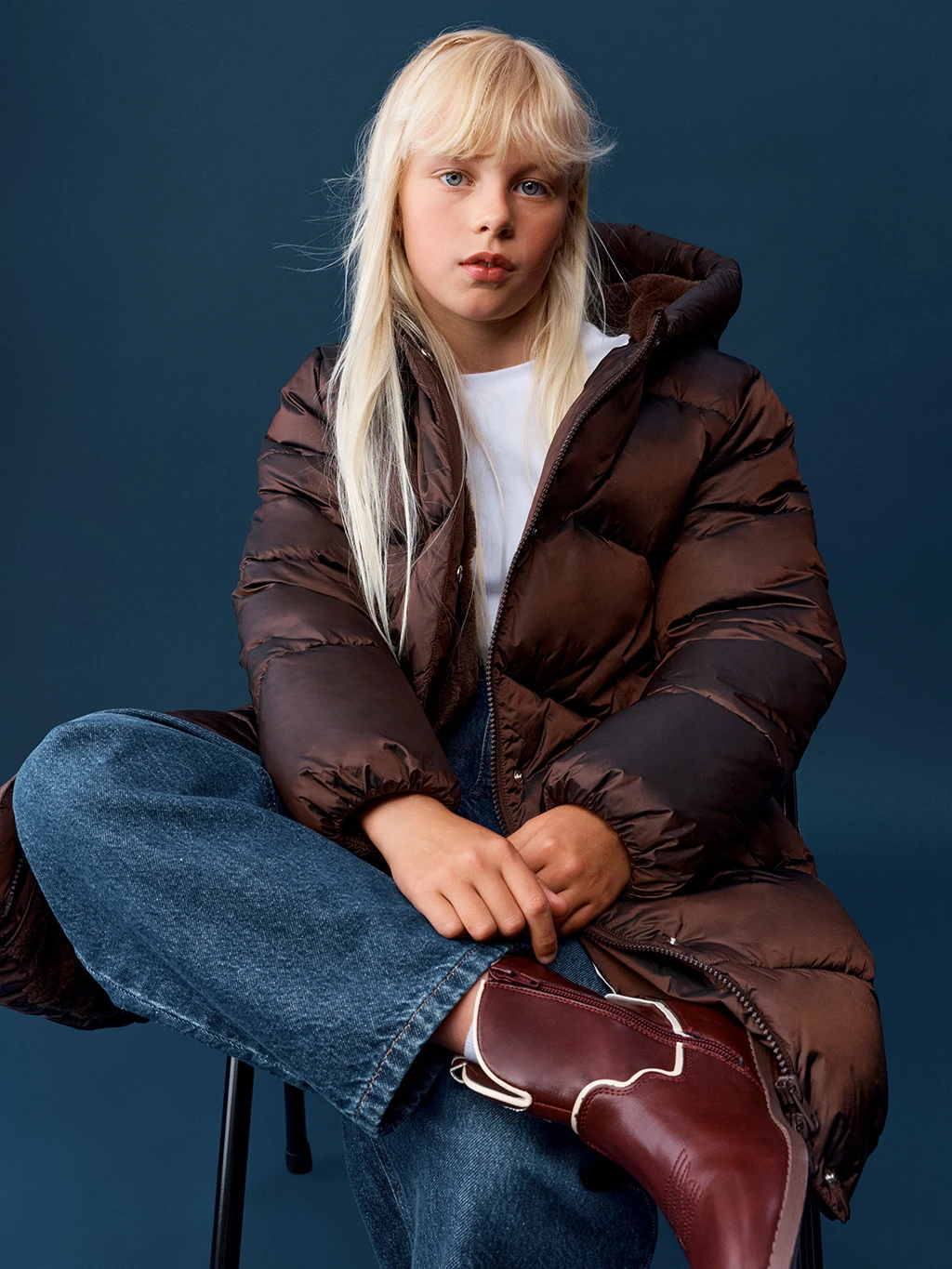 Young child in a brown long puffer jacket, wide leg jeans and red cowboy boots.