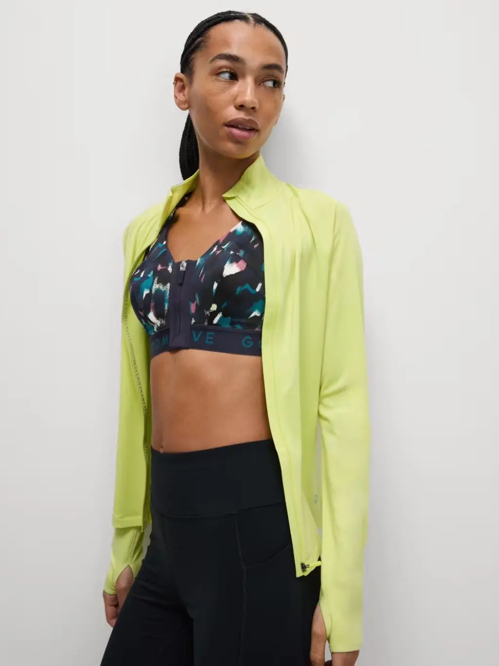 A woman wears a multi-coloured sports bra under a bright lime green sports jacket and with black leggings