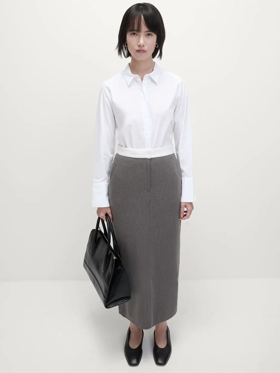 Woman wearing a grey pencil skirt with a white skirt, black ballet pumps and a black bag.