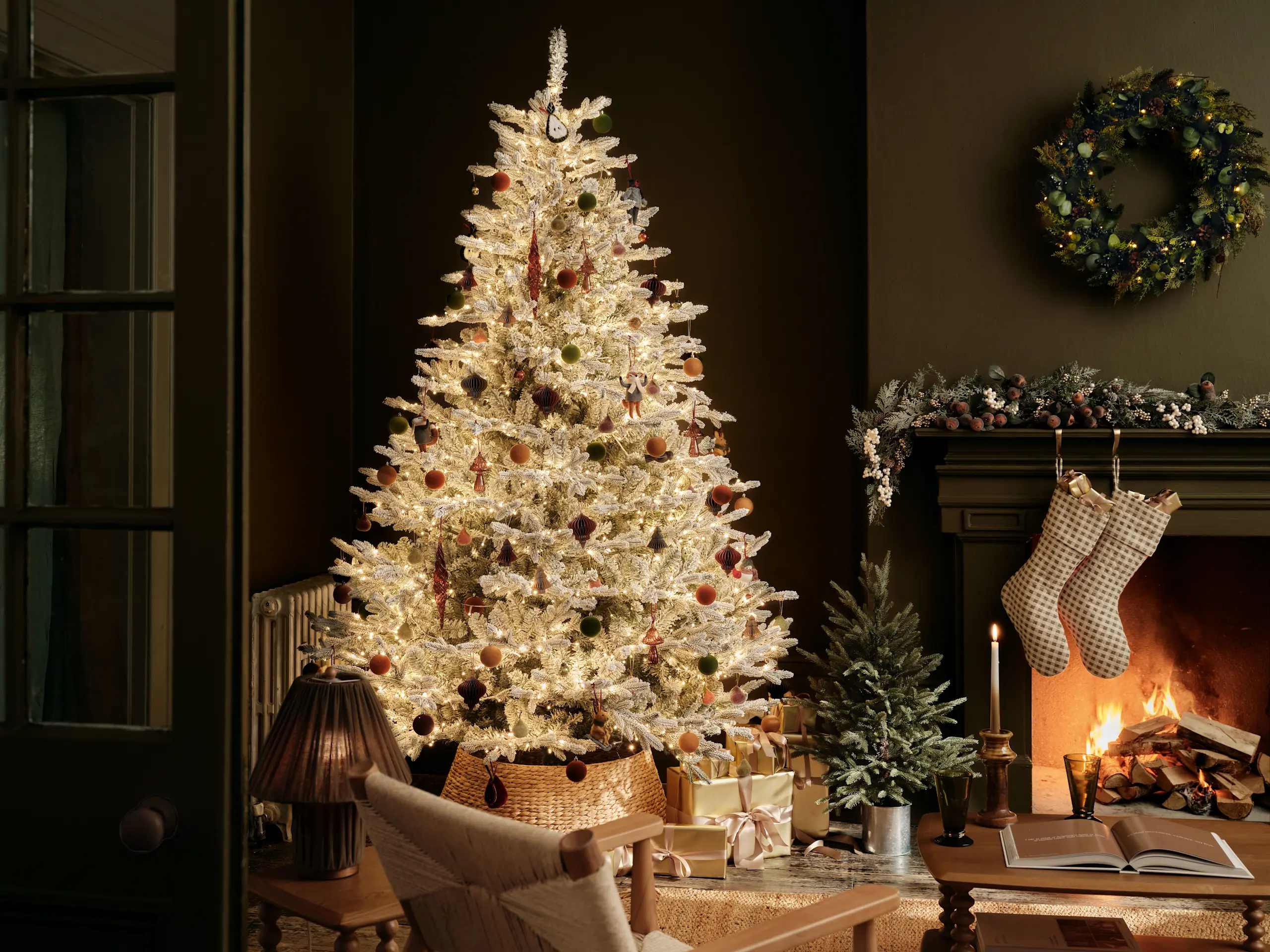 White Christmas tree next to a mantelpiece, stockings and evergreen foliage