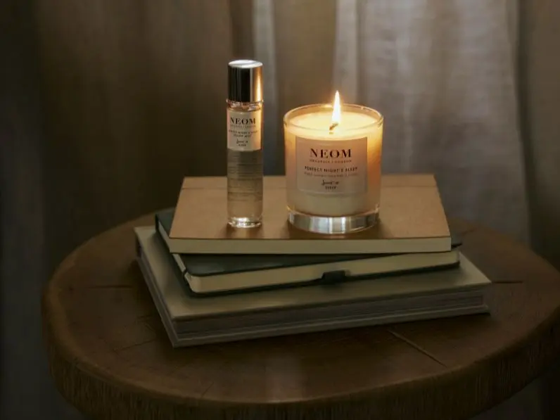A candle and bottle of skincare sit on a bedside table on top of some books with a drawn curtain behind