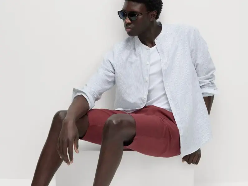A man, sitting down, wears burgundy shorts and striped shirt