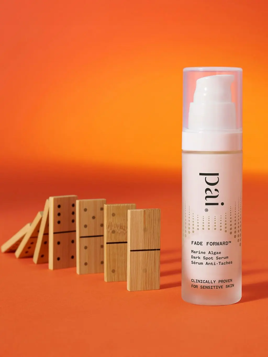 A shot of Pai Dark Spot serum against an orange background next to a row of wooden dominoes