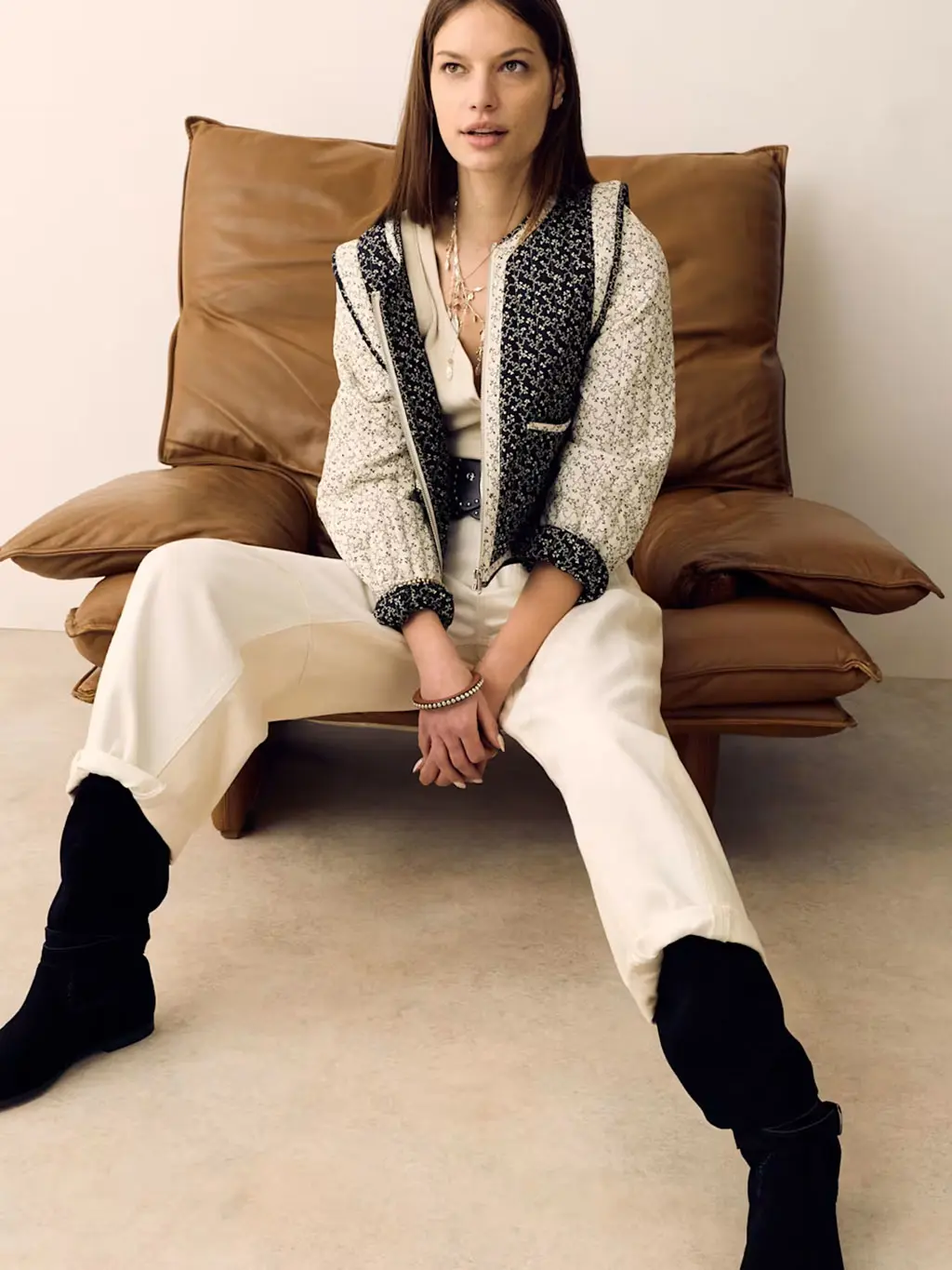 Woman wearing a cream vest and jeans with an oversized studded belt, quilted jacket and black slouchy boots