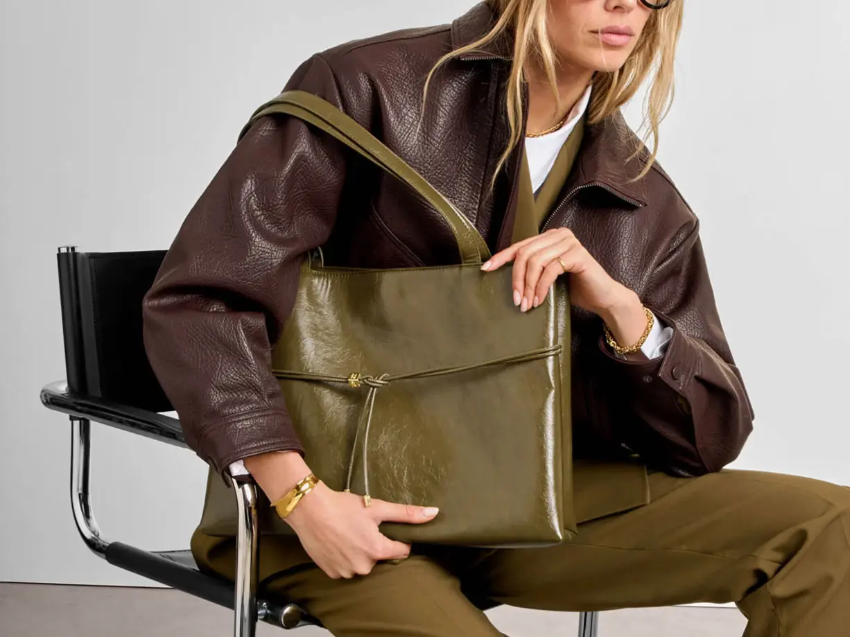 Woman wearing brown leather jacket and olive-toned trousers, holding a matching leather olive handbag 