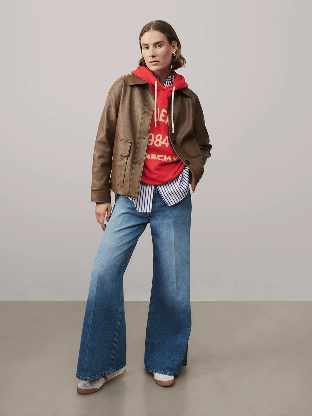 Woman wearing brown jacket, red hoodie, blue jeans and red and white trainers