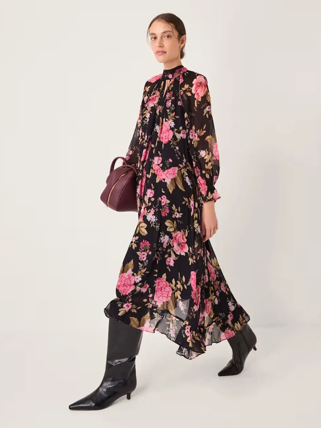 A woman wears a dark floral ruffle dress, black boots and carries a burgundy handbag