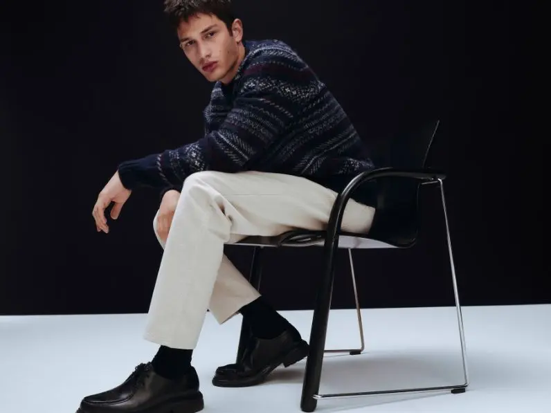 A man sits on a chair wearing cream jeans, black shoes and a blue fairisle jumper