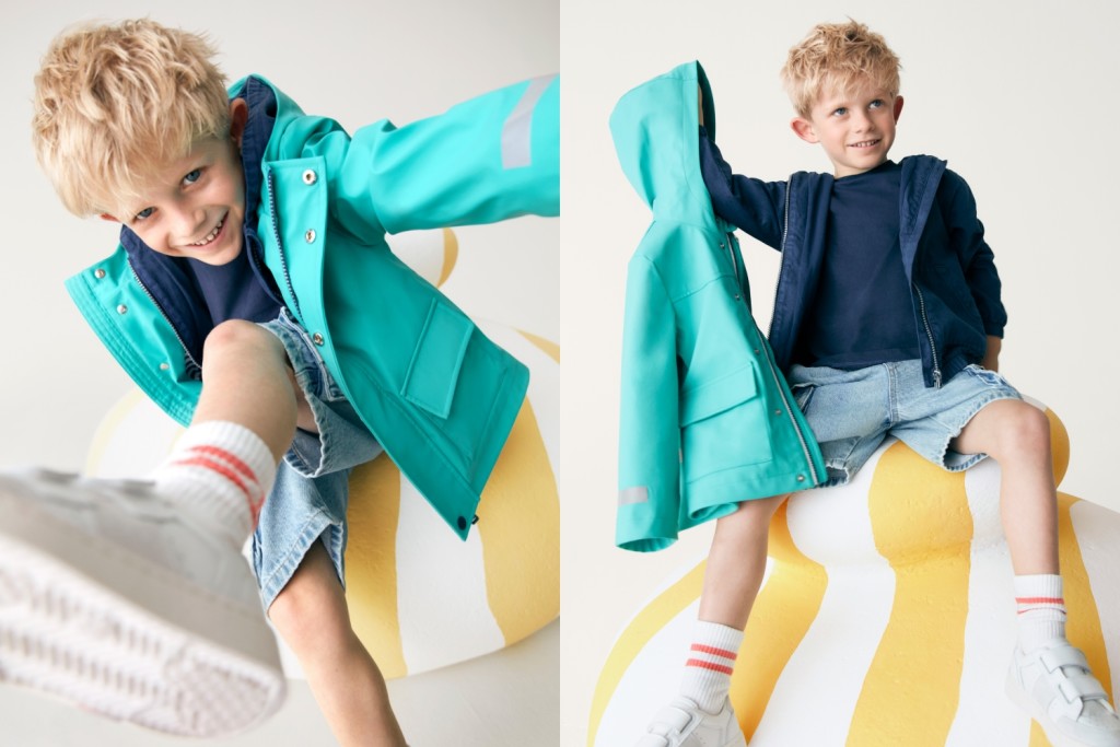 Boy wearing a teal raincoat over a navy top and matching hoody with denim shorts and white trainers. Shop raincoats