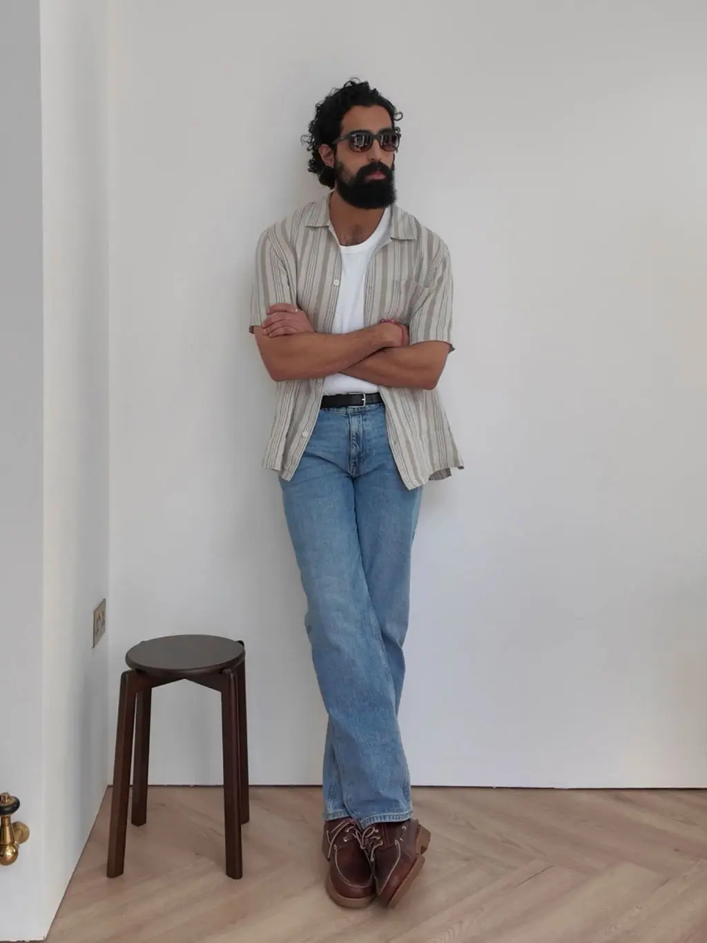 Tom Cheema wearing a white vest, striped shirt, blue jeans, brown boat shoes, black belt and sunglasses