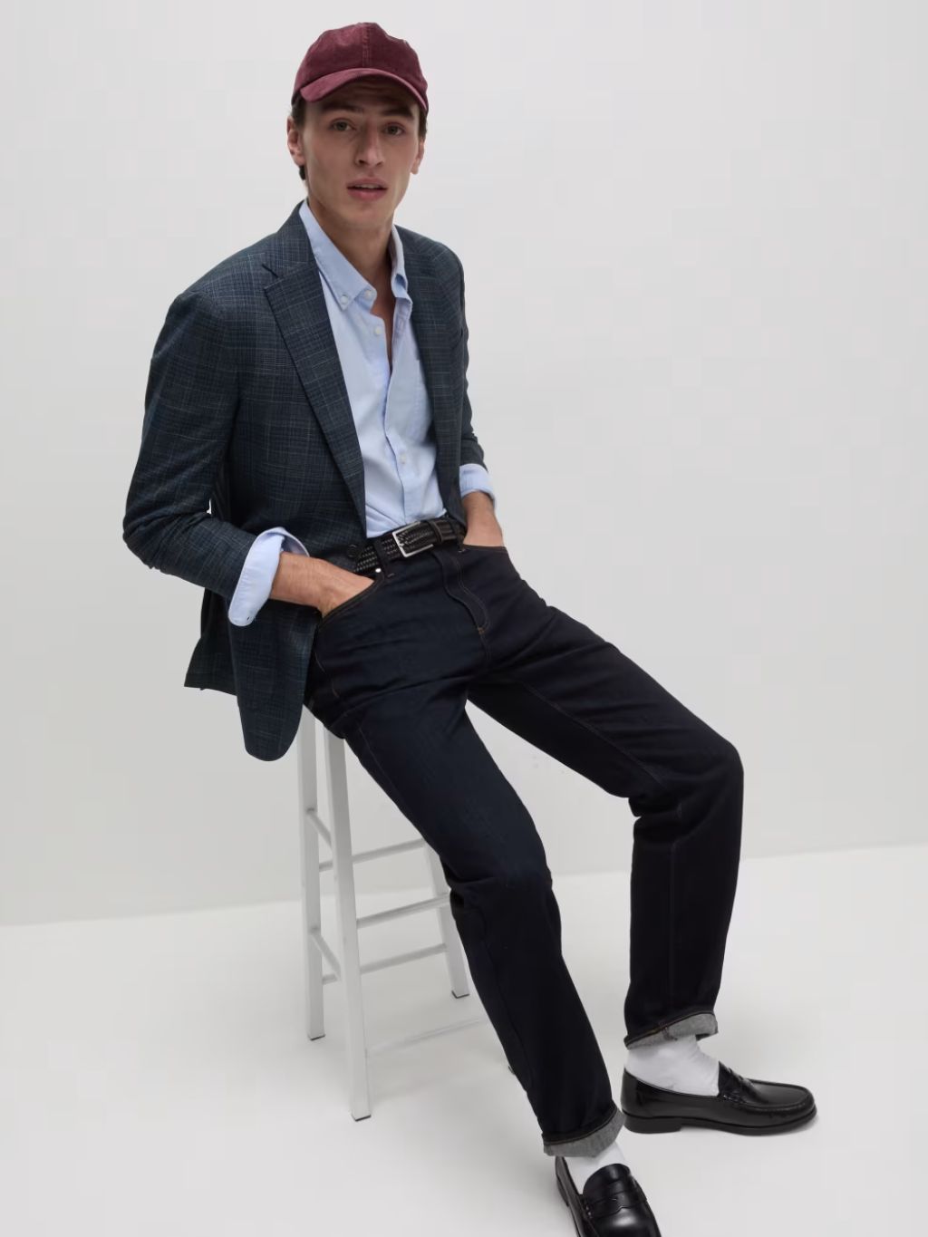 A man sat on a stool wearing a blazer, oxford shirt, jeans, loafers and a baseball cap