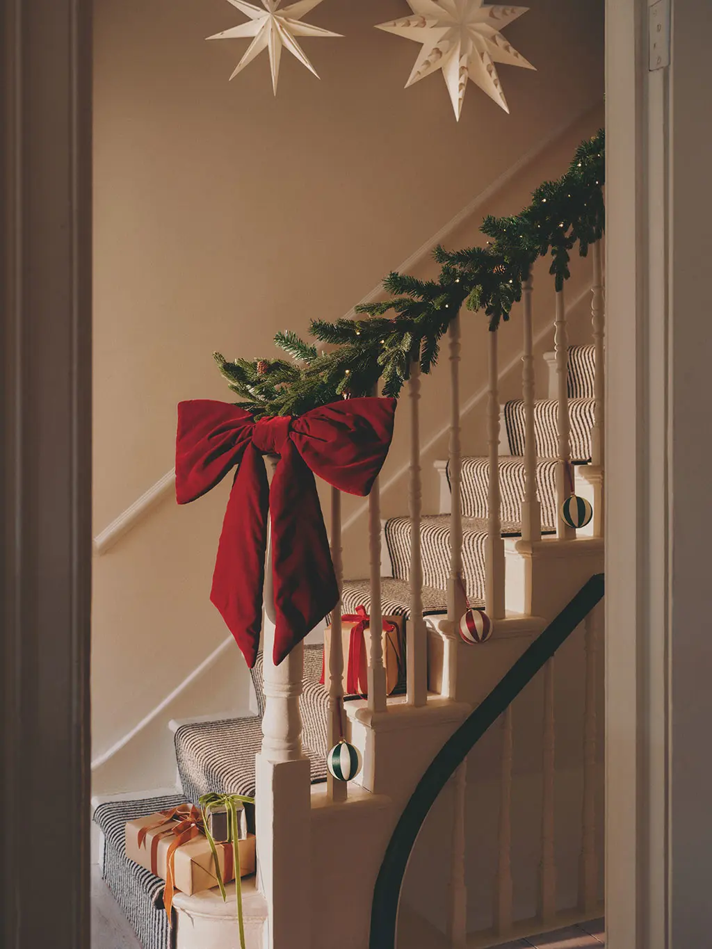 Cream staircase with a green garland wrapper round the banister, large red bow and presents along the bottom of the stairs.