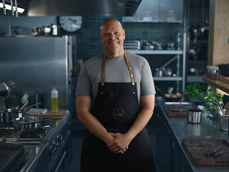 Tom Kerridge wearing a grey T-shirt and an apron in a kitchen