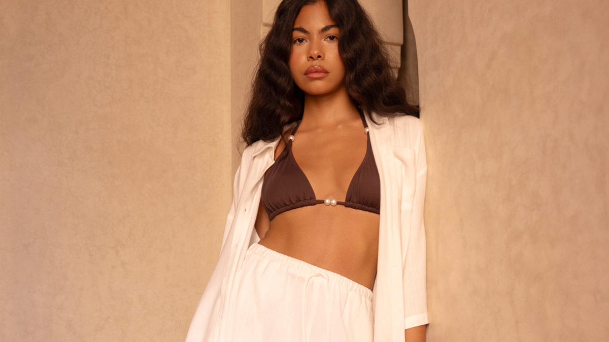 Woman wearing a brown bikini top, white beach shirt and white linen trousers