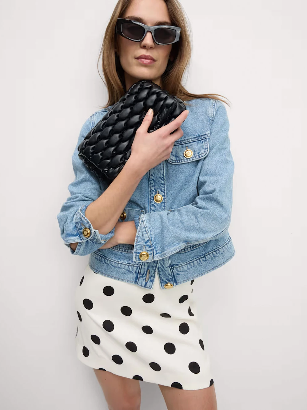 Woman wearing sunglasses, a blue denim jacket, a black and white polka-dot mini skirt and a black quilted clutch