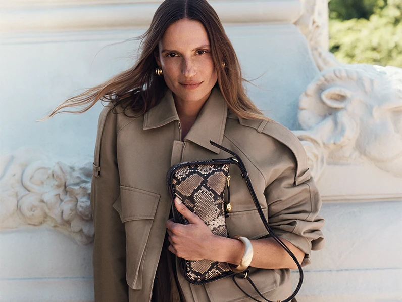 Woman wearing light brown jacket holding a snake-print shoulder bag
