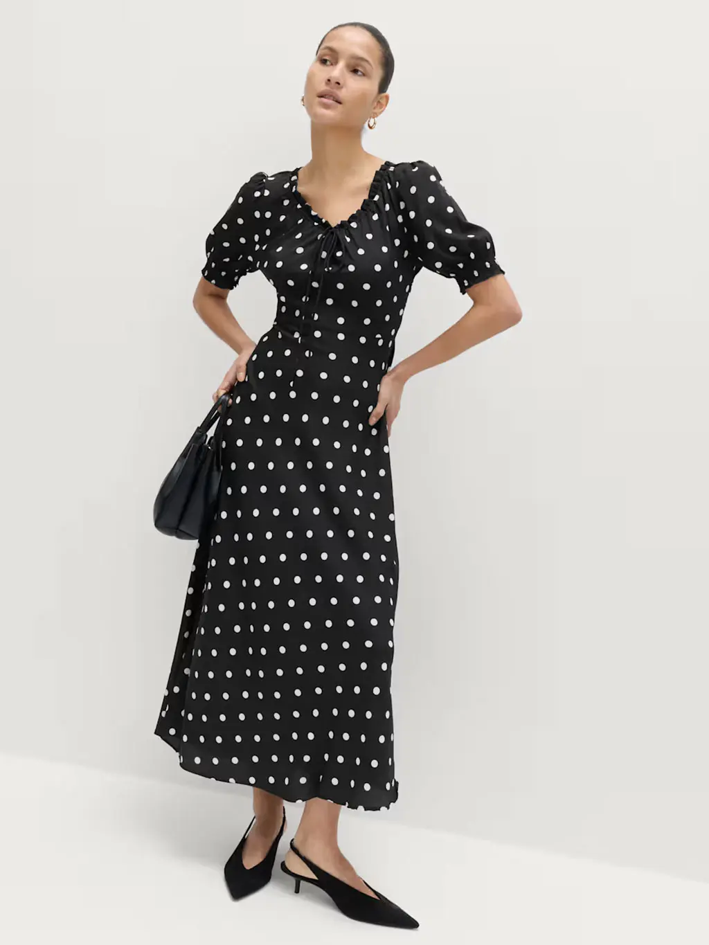 Woman wearing polka-dot dress with gold hoop earrings, a black bag and black kitten heels