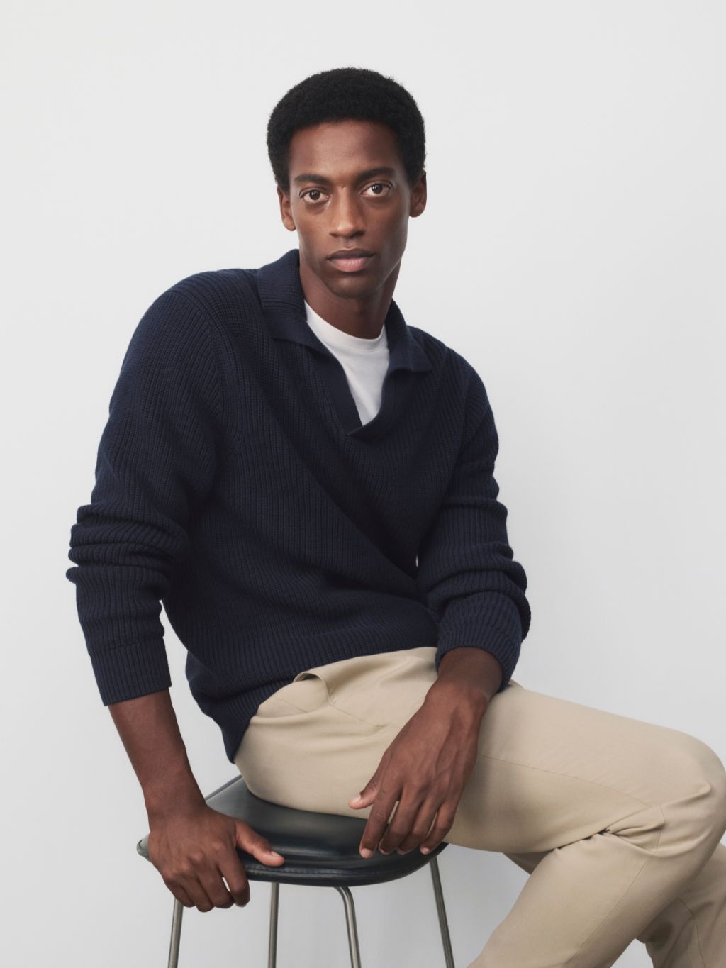 A man sat on a stool staring at the camera wearing a navy jumper, white t-shirt and chinos