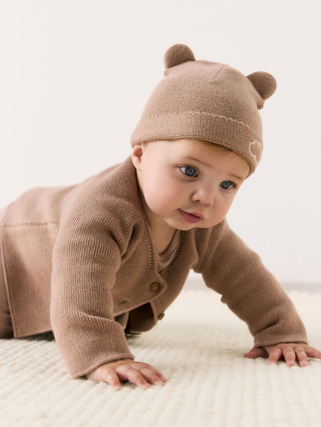 A baby is crawling on the floor wearing a brown outfit including a hat with ears