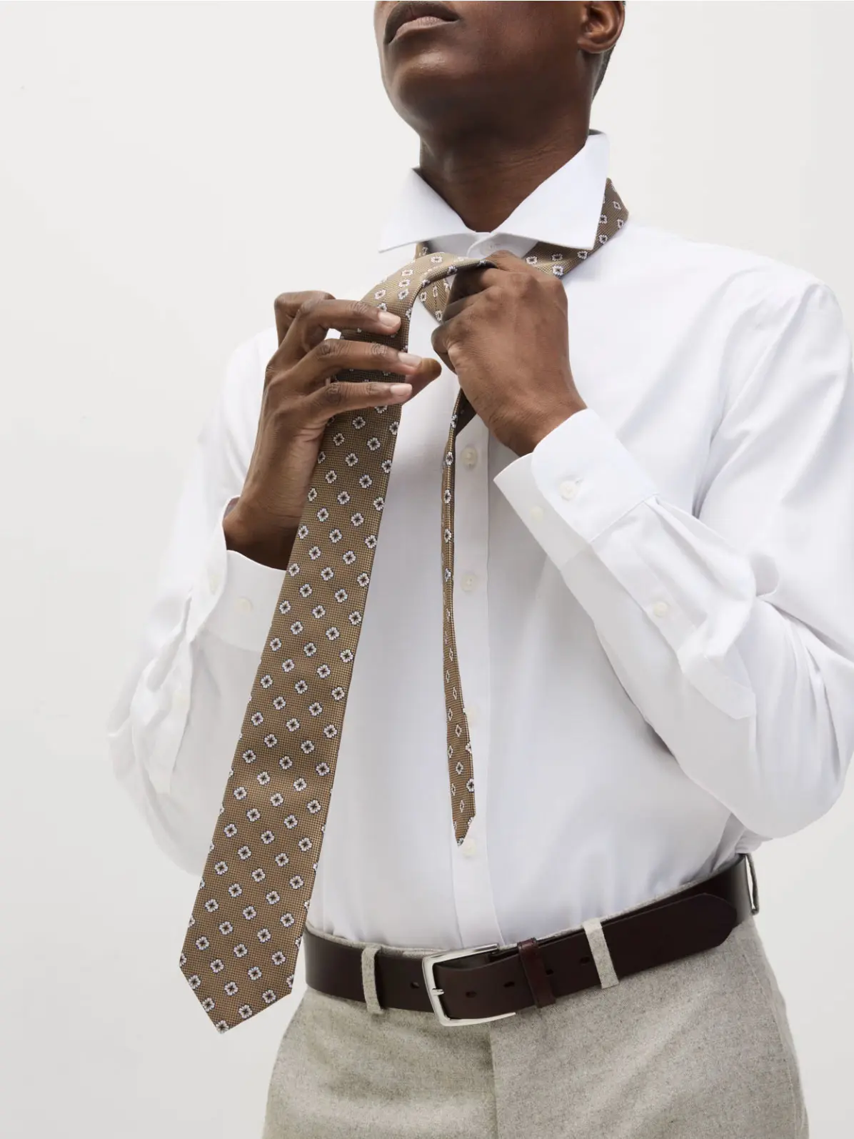 Man wearing white shirt, neutral trousers and a belt, tying a brown patterned tie