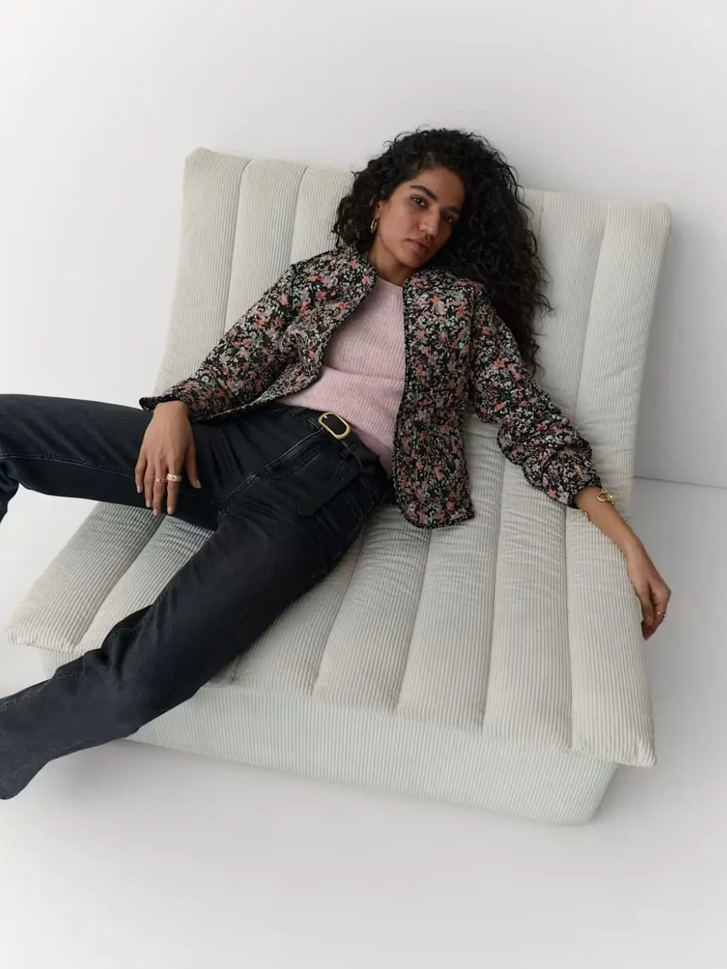 A woman lies on a chair wearing a Per Una floral quilted jacket, pink jumper and black jeans
