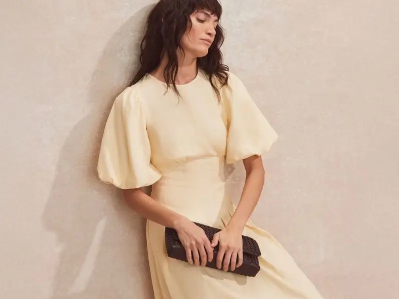 A woman wears a pale yellow linen dress and carries a dark clutch bag