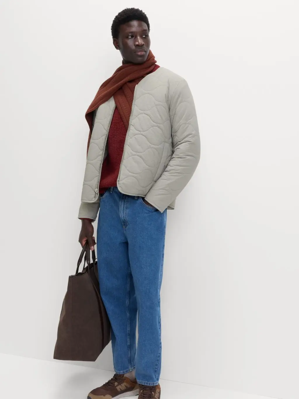 A man wears a quilted jacket, scarf and jeans, carrying a tote bag 