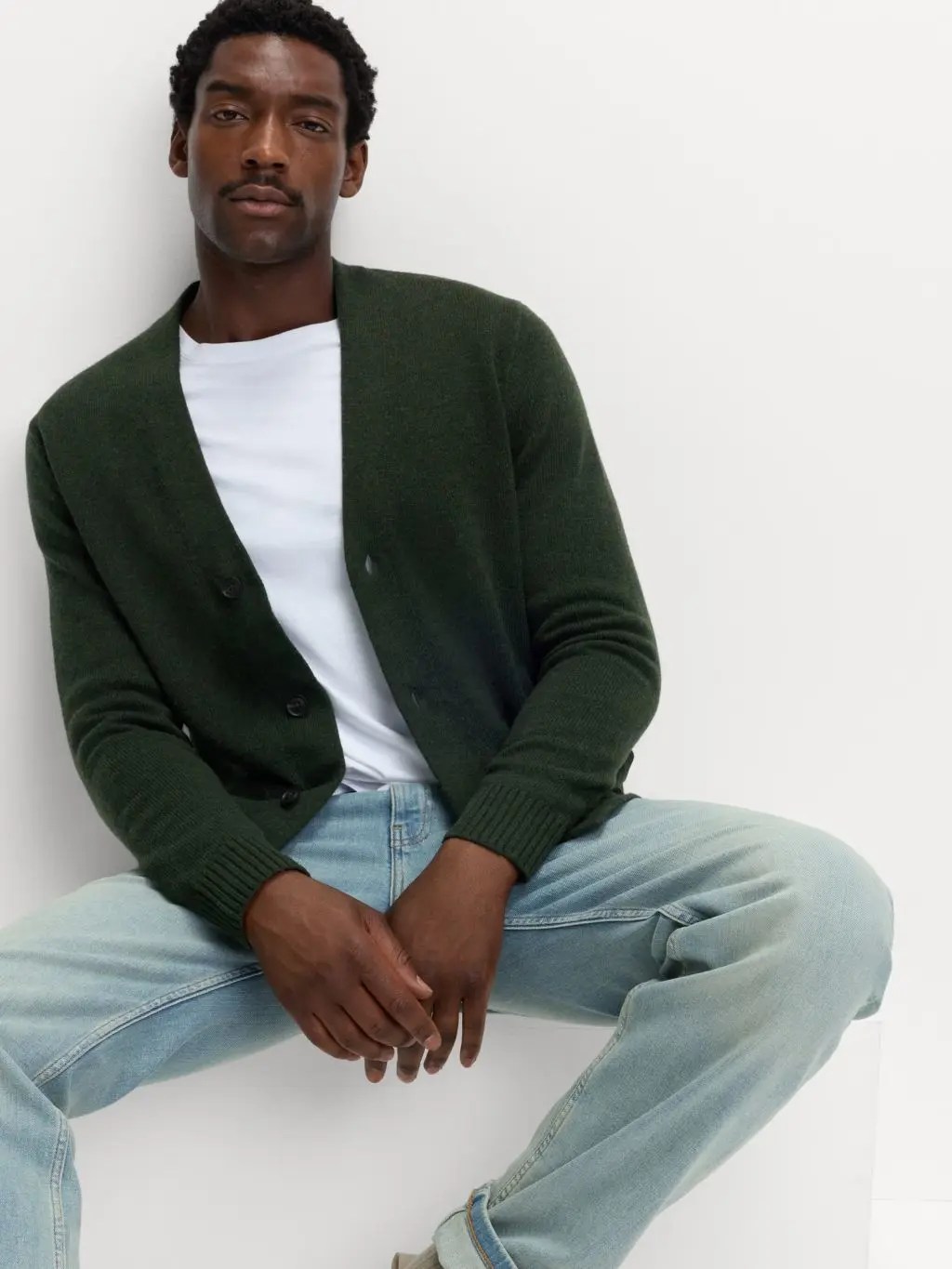 A man is sat down wearing a khaki cardigan over a white t-shirt with pale jeans