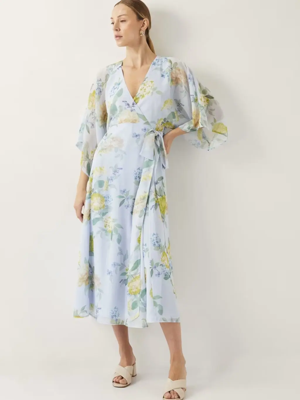 A model wearing a wrap dress with a pretty blue, green and yellow floral print, stood against a pale wall