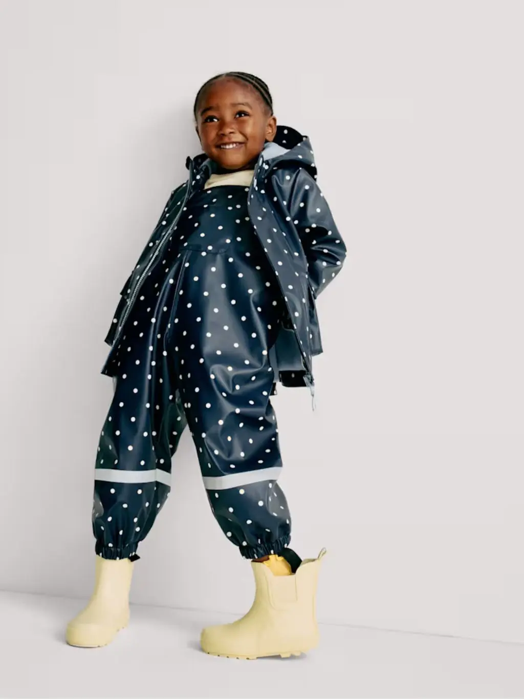 A young girl leans against a wall wearing waterproof dungarees, raincoat and yellow wellies