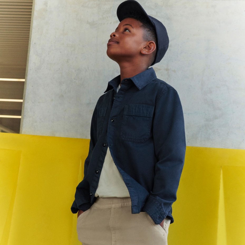 Boy wearing a black shirt over a white top with beige chinos and a black cap. Shop boys’