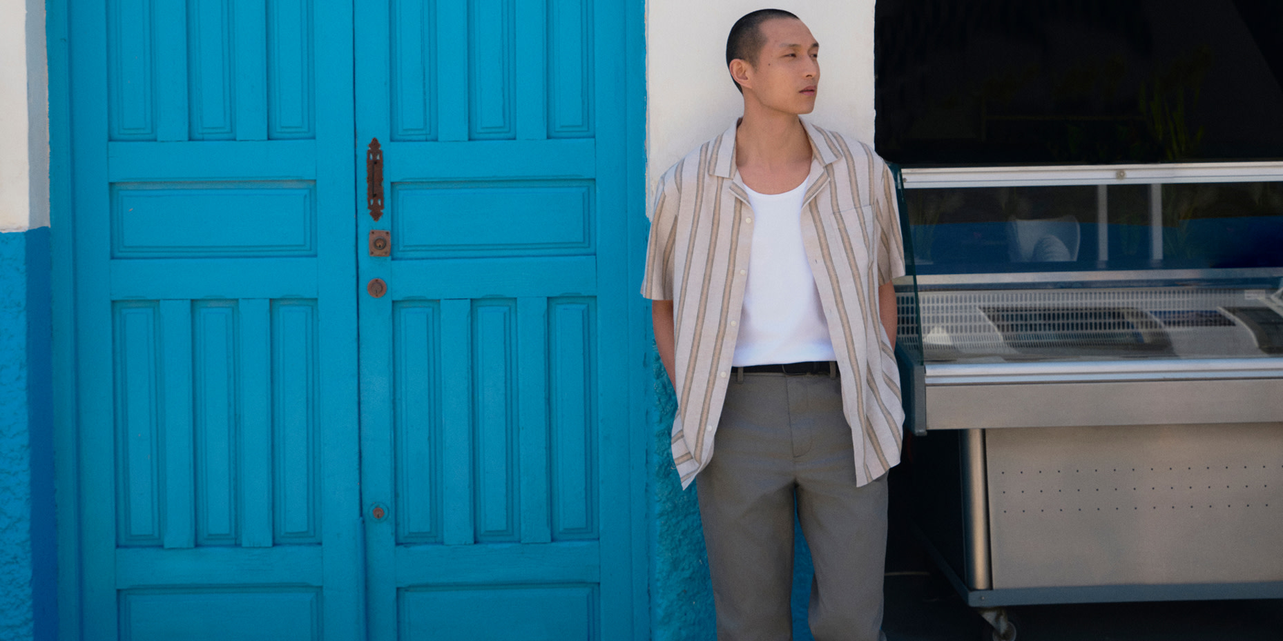 Man wearing a striped shirt, white T-shirt and grey trousers beside a pair of blue doors