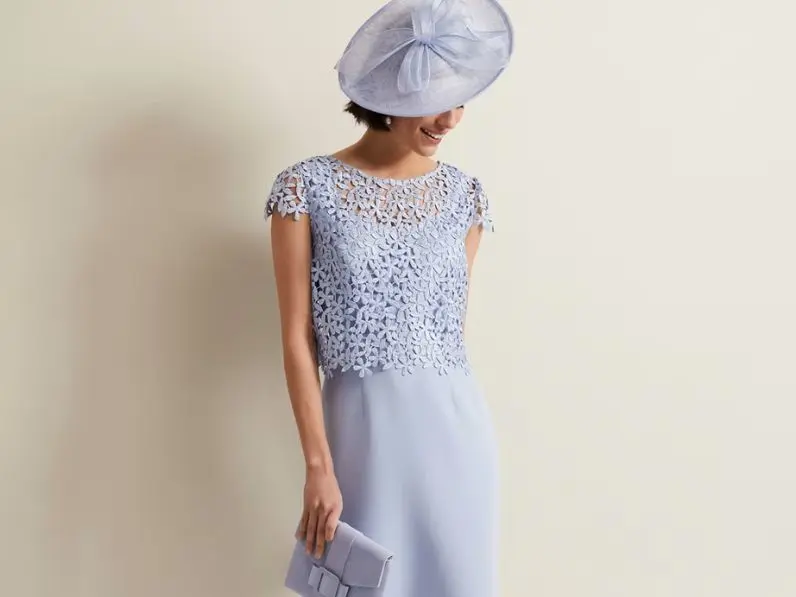 A woman wearing a lilac fascinator and matching dress stands against a pale cream background