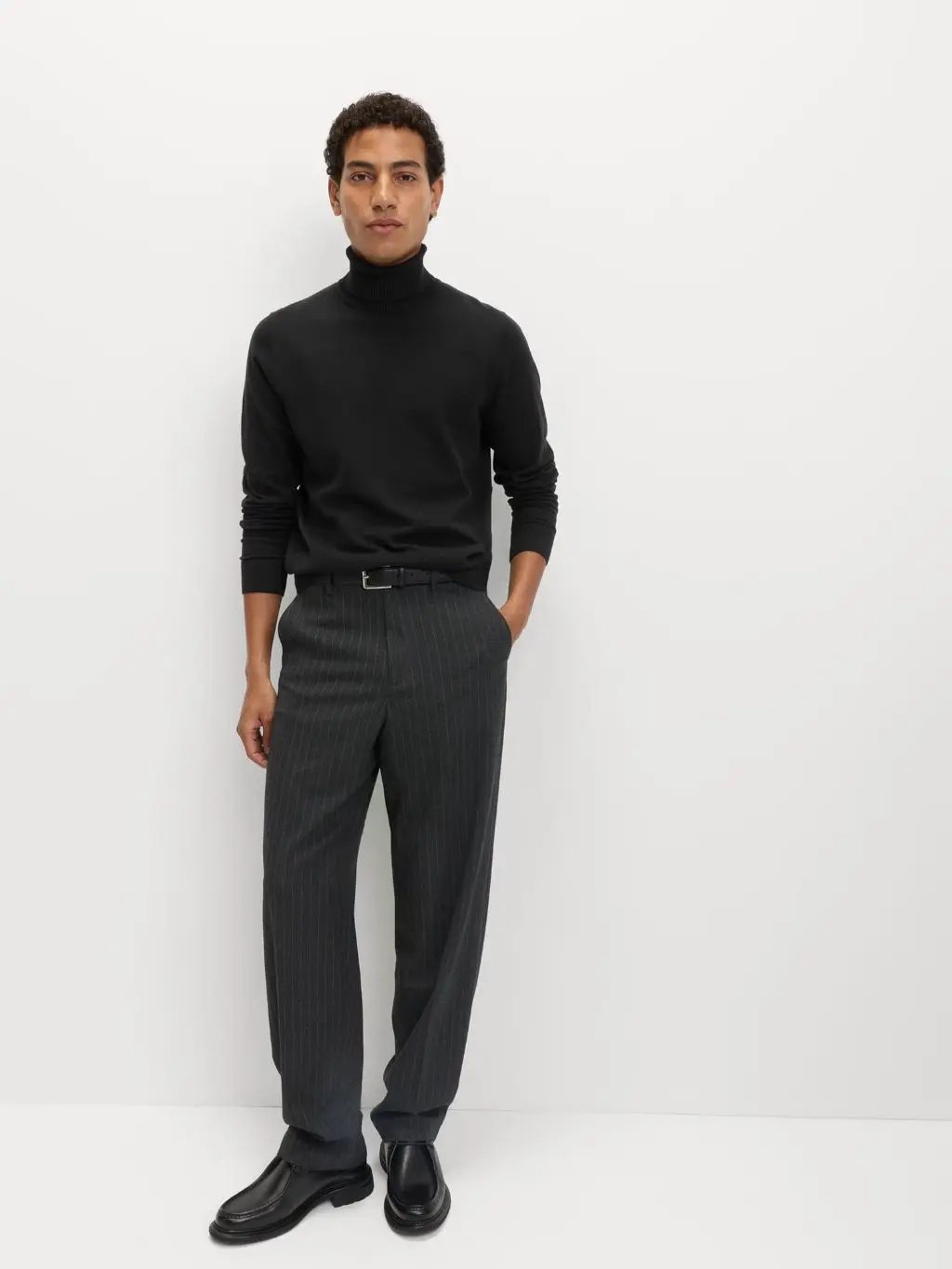 A man leans against a wall wearing a black roll neck and grey trousers