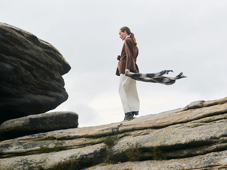 Woman standing on a rock face wearing white trousers and a brown jacket and holding a slim checked scarf.