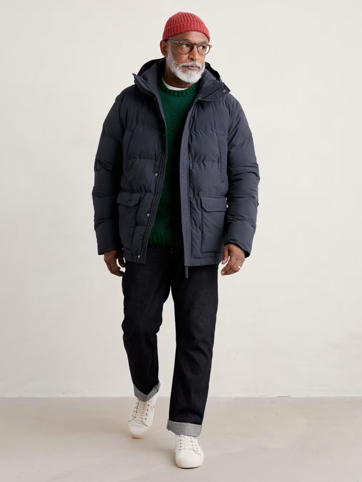 A man in glasses, wearing a red beanie hat, navy puffer coat and dark jeans and trainers