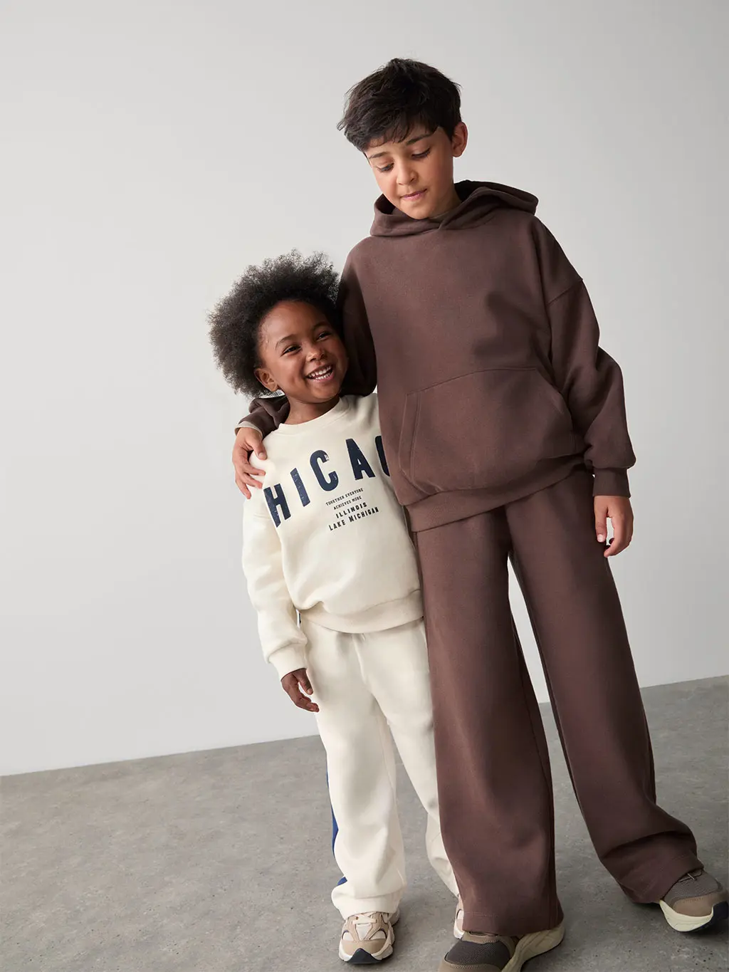 Boy in Chicago-print sweatshirt and joggers with trainers beside a boy in a brown hoodie, joggers and trainers