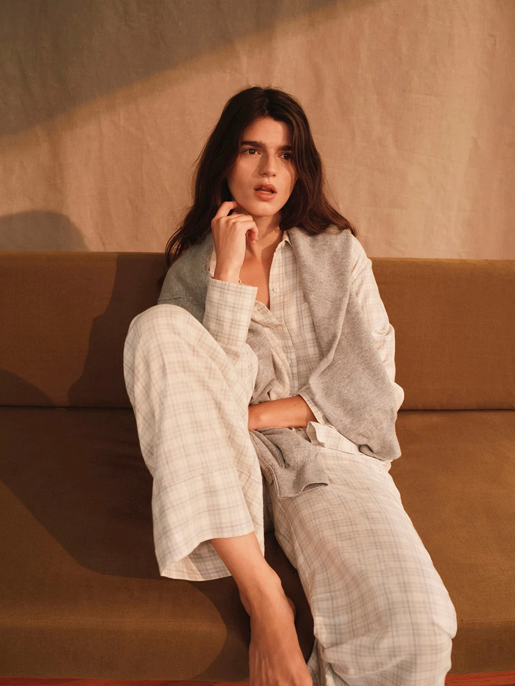 Woman wearing a grey checked long pyjama set sat on a brown sofa.