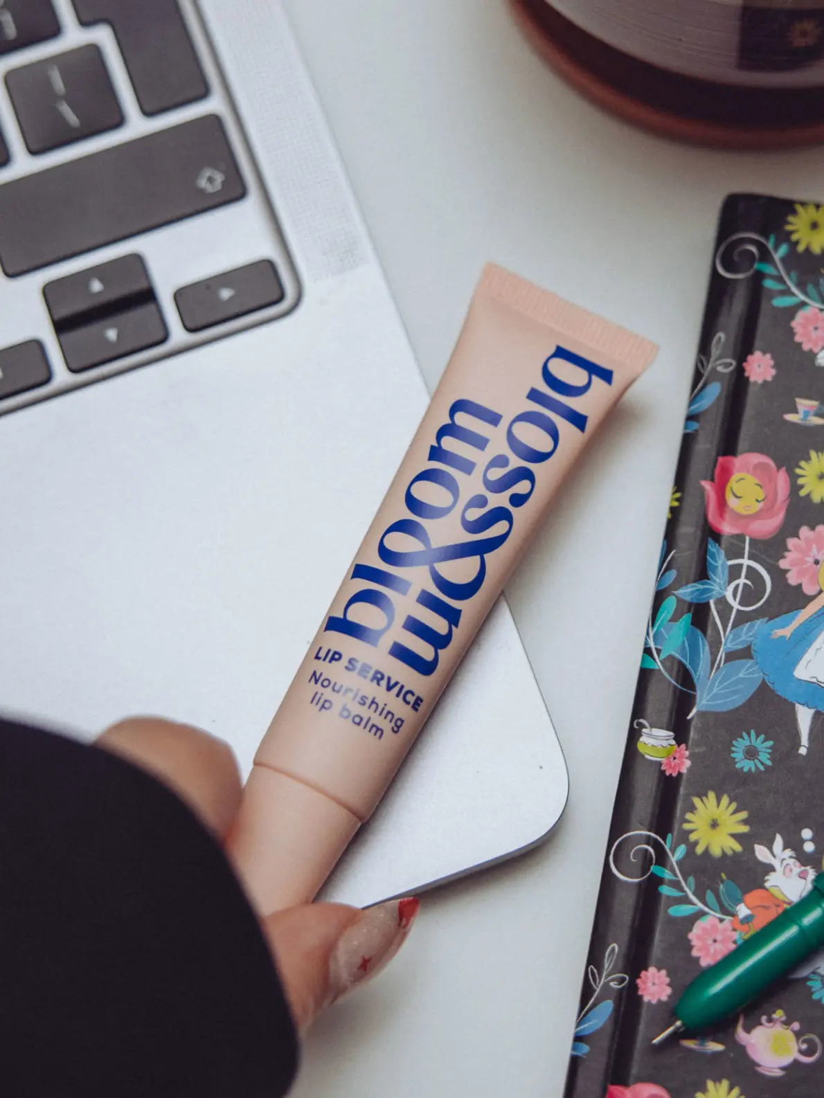 Woman's hand holding tube of lip balm in baby pink and blue beside open laptop and notebook