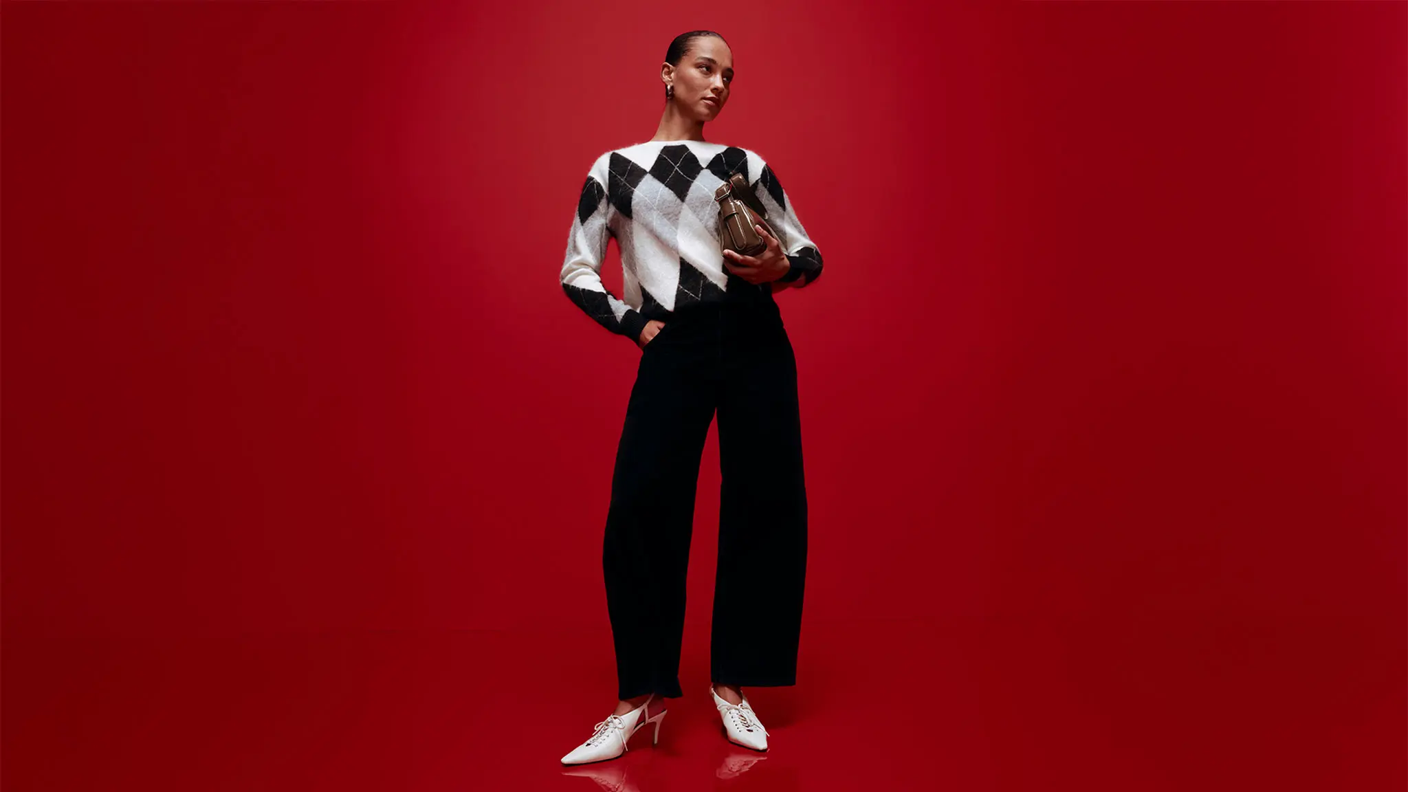 Woman wearing black barrel-leg jeans with an argyle jumper, white heels and a clutch bag