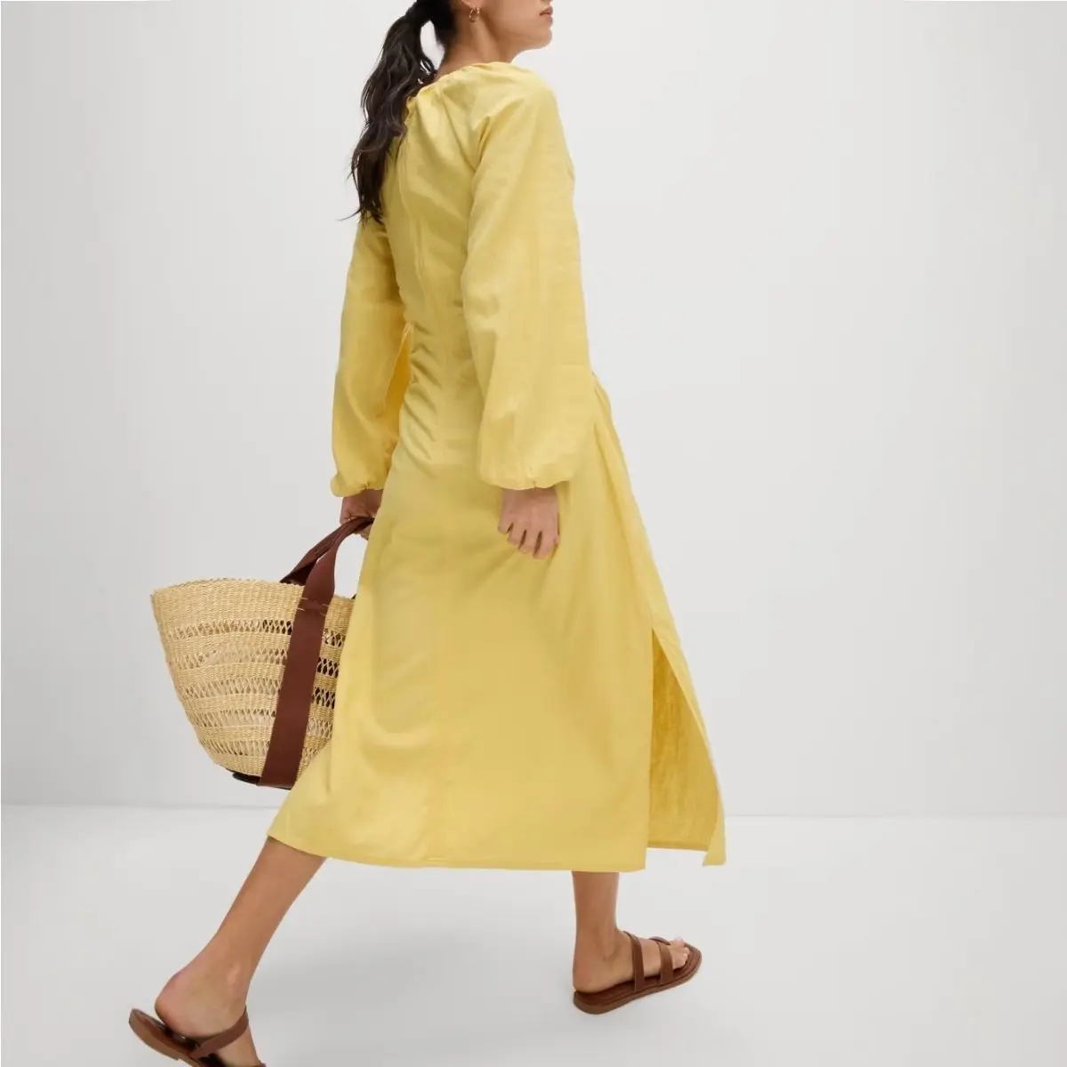 A woman wears a butter yellow linen dress and sandals, carrying a straw tote bag