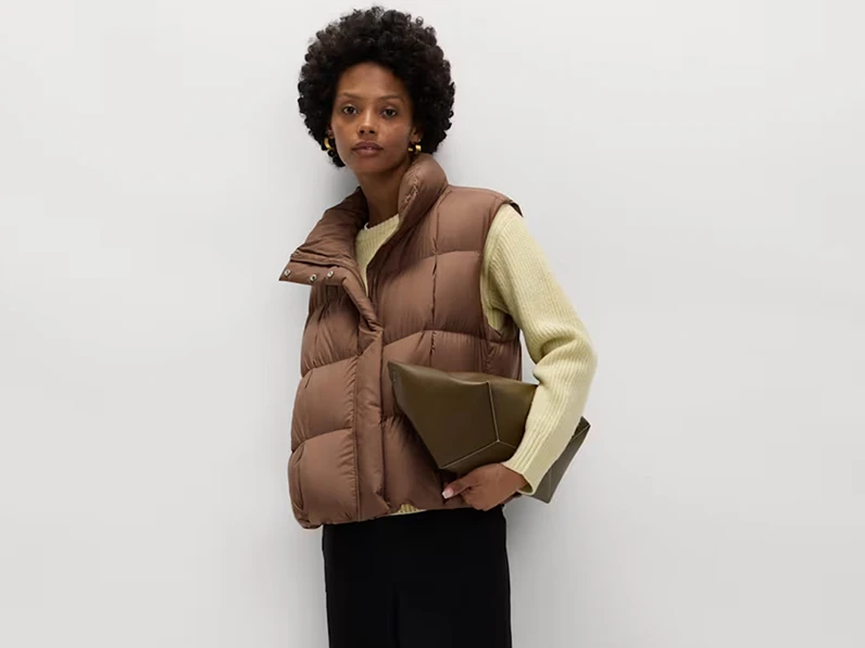Woman wearing a brown puffer gilet, green jumper and black trousers.
