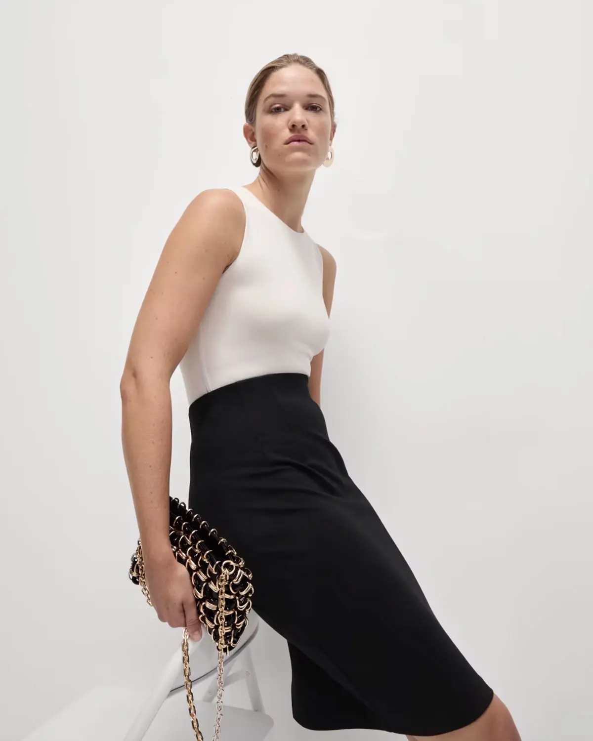 Woman wearing a white vest tucked into a black midi skirt with a black bag and gold earrings