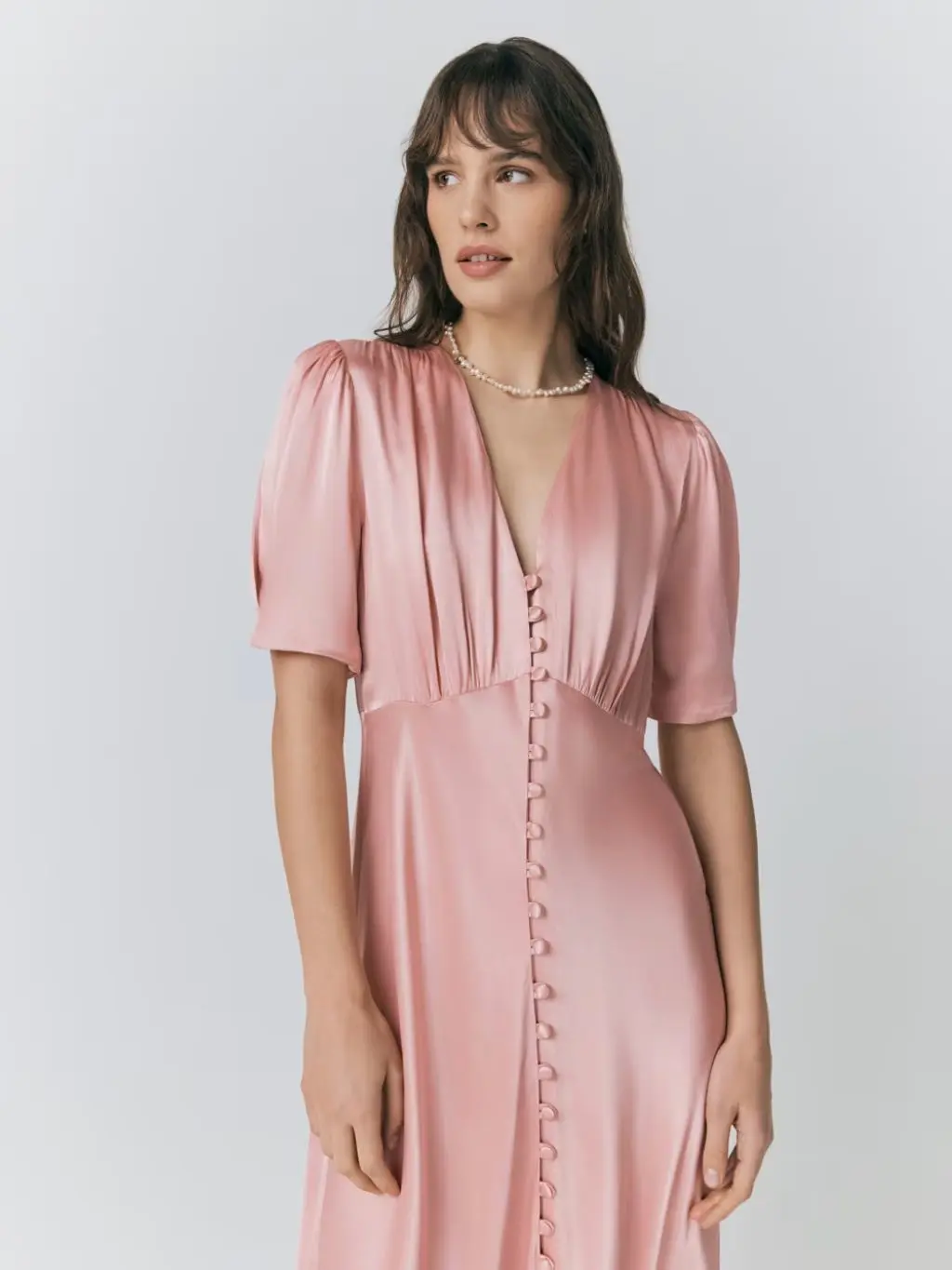 A model, looking away from the camera, wears a pink satin dress and silver necklace