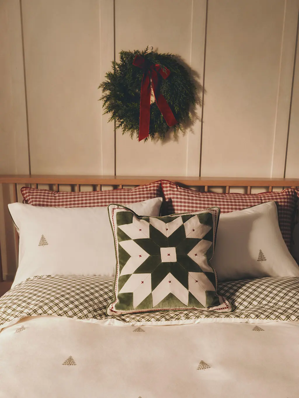Wooden bed with green and white bedding, patchwork cushion and wreath with a red ribbon hung above.
